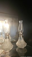 Pair of two clear glass oil lamps with ribbed textured bases and silver-tone burner assemblies, shown against a mirror background.