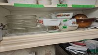 Shelf with stacked clear glass bakeware, white Pyrex and FireKing casseroles with floral and fruit patterns, and nested brown-toned Pyrex Old Orchard bowls.