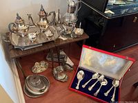 Photo showing full silver plate tea and coffee set displayed on tables including tray, coffee pot, teapot, cream and sugar, additional serving pieces and a set of spoons in a red case.