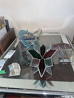 Five stained glass pieces displayed on a glass table including starburst, heart-shaped atom, leaf, candle holder and trident designs.
