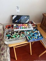 Multiple containers of thread spools displayed on side table; two clear bins, one with greens/blues, one with neutrals/pastels, plus a Janome branded box and a tub of reds/purples