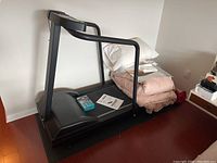 Black treadmill viewed from side with owner's manual and some folded linens on deck. Mat underneath treadmill.