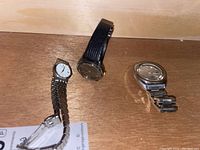 Three wristwatches on a wooden surface: Citizen Quartz with two-tone metal band and two Seiko quartz watches, one on black leather strap, one on silver metal bracelet.