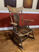 Side angled view of the antique wooden rocking chair showing the curved backrest with a wide top rail and solid wood seat with worn finish.