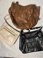 Three purses grouped on a light surface: beige rectangular with front zipper, brown soft leather slouchy purse, black leather handbag with front zippers