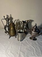 Five pitchers arranged on a surface showing different sizes and designs including both silver plated and pewter materials.