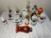 Overview of 13 miniature oil lamps of various designs and colors on white cloth background.