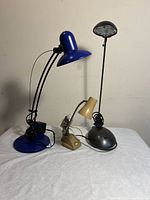 Three table lamps displayed on a white cloth against a wall: blue adjustable lamp with dome shade, beige small lamp with curved neck, black tall lamp with circular element near neck.