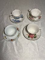 Four tea cups with matching saucers laid out on a white cloth background showing different floral patterns and gold trims.