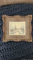 Overall view of the framed art print on a textured blue surface showing the detailed ornate frame and the small size of the print.