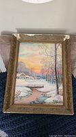 Frontal view of oil winter landscape painting showing snow, trees, stream, houses, and sunset sky in detailed ornate frame.