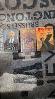 Three hardcover books displayed on a carpet, including two Harry Potter books and one autographed Colin Powell book.