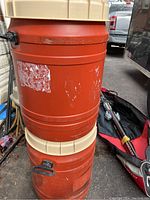 Two stacked orange plastic barrels with beige screw caps and black handles, showing some wear and label residue.