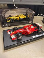 Yellow Mugen Honda Formula 1 car in original Hot Wheels box displayed behind the red Ferrari F1 model car on a black stand.
