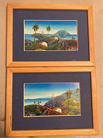 Pair of framed miniature landscape paintings with coastal hill scenes, palm trees, and small houses, in wood frames with blue mats.