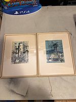 Photo showing both framed lithograph prints from a distance, placed side by side on a surface
