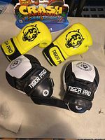 Top view of yellow Tiger Pro boxing gloves and black and white training mitts on a surface with Crash Bandicoot display in background.