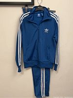 Front view of the blue Adidas Originals track jacket hanging with matching pants visible below
