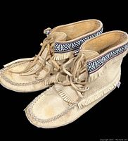 Side angle of lightly worn tan leather moccasin boots with concho trim and fringe detailing.