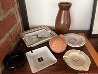 Photo showing five ashtrays, wooden jug, and terracotta ornament arranged on a wooden surface.
