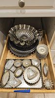 Top-down photo showing a wood drawer containing stacked metal fluted tart pans and various small metal molds of different shapes including rectangular, oval, heart, and fluted designs.