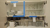 Full view of cabinet with white ceramic plates stacked, white ceramic pitcher with metal lid, dark brown coffee cups with 'Cappuccino' text, clear glass pitcher and multiple clear glasses with handles.