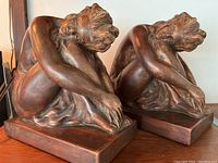 Pair of seated female nude metal figurine bookends on a wooden surface, showing front and side views.