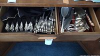 Wooden drawer with rows of ornate silver-plated forks and a mix of serving utensils in adjacent compartment