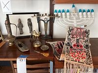 Wide shot showing the brass menorah, electric menorah with blue bulbs, brass containers including camel, ewer, shoe, small tray, and a pair of framed carved figures; two fabric pieces with Judaic tapestry designs with hamsa patterns.
