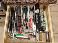 Top view of kitchen drawer showing a wooden rolling pin, multiple knives and blades, a metal handheld grater, knife sharpener, and various spoons.