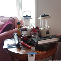 Image showing a round wooden table with two vintage style battery lanterns with beige bases and blue tops, a red compact camping lantern, multiple flashlights in different colors and sizes, and a black fluorescent battery-operated light with amber indicator. Also includes a sealed pack of replacement fluorescent bulbs.