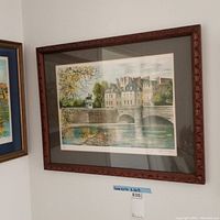 Framed signed wall art against a corner wall showing a cityscape with a stone bridge, water reflection, trees, and a statue. 20x25 inch frame, 13x19 inch art.