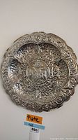 Front view of round silver-plated wall plaque with embossed heraldic crest and ornate scroll design