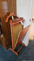 Valet stand view showing upright folded posture with hanger and trouser press panel facing forward against a wooden cabinet background.