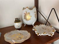 Four vintage French porcelain pieces: two dishes, one vase, and a table lighter displayed on a wooden shelf, showing ornate gold hand-painted floral designs.