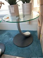 The glass top shows the round, clear surface held by the curved silver metal base mounted on a black round platform on blue carpet.