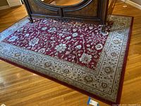Photo showing the full area rug underneath a piece of wooden furniture on a hardwood floor, highlighting the red field and cream border with floral and geometric patterns.