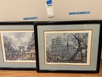 Two framed prints resting against a wall, one showing Notre Dame cathedral with a tree and people, the other showing Arc de Triomphe and a street scene with cafes, including 'CAFÉ PARIS' signage. Both frames tallied with measurements taped above.