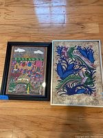 Frontal view of both framed paintings placed on wooden floor showing vibrant folk art scenes on bark paper.