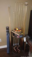 Photo showing nine wooden pool cues standing in a wooden rack stand, two soft cases for cues leaning against wall nearby, wicker basket with pool balls on the rack, and other accessories scattered around.