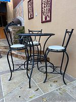 Overall view of wrought iron bistro table and three chairs with cushions arranged on a tiled patio by a pool.