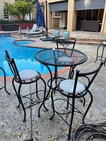 Full view of wrought iron bistro set by pool including 3 chairs with white cushions and round mesh table.