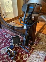 Full view of SlimCycle upright exercise bike with seat and handlebars. On floor near bike are multiple dumbbells, an ab roller wheel, and the Med massager showing cords.