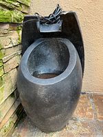 Front-on photo showing the black stone electric fountain with power cord on top, placed against stone wall and beige background.