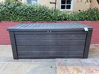 Front view of the dark plastic patio storage box with faux wood paneling and hinged lid on outdoor tile.