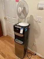 Long shot of Brio water dispenser with Atlantic Breeze fan mounted on top, against wall with light switch and wooden floor.