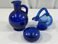 Photo showing set of three blue glass decorative items including vinegar bottle, small pitcher, and round vase