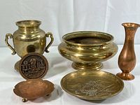 Six items including a polished brass urn with handles, a plain brass bowl, a brass souvenir medallion from Singapore, a small copper bowl, a copper bud vase, and a decorative etched brass bowl.