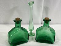Front view of two matching green glass bottles with cork stoppers alongside a tall slender green glass bud vase on a white background.
