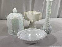 View of all four milk white glass items including a lidded canister with embossed grapes, a square candy dish on pedestal, an oval dish with beaded edge, and a tall bud vase with quilted pattern.